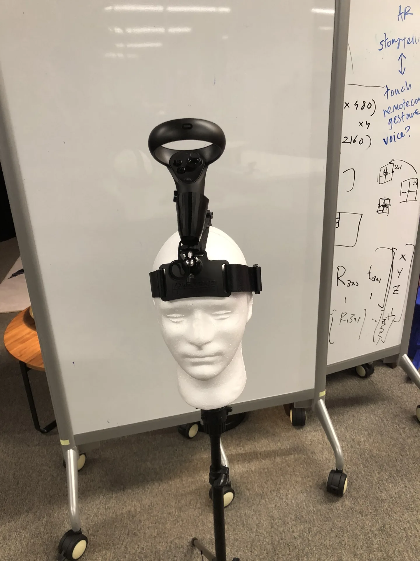 A white foam mannequin head on a stand wearing a strap with a VR controller mounted on top of it, in front of a whiteboard.